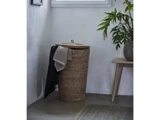 En høj flettet vasketøjskurv med låg står i et hjørne med et beige og et sort håndklæde. I nærheden står et træbord med en potteplante, og der kommer sollys ind gennem et vindue ovenover.