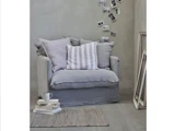 En hyggelig grå lænestol med stribede puder står ved en stak bøger og en kop ved siden af et tæppe. Indrammede fotos hænger på væggen over stolen i et minimalistisk rum.