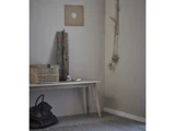 Et minimalistisk rum med en træbænk, en flettet kasse, drivtømmer, sten, en sort taske og væghængt rustik indretning mod en lysegrå væg.