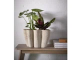 En struktureret beige krukke med grønne og lilla stueplanter står på et træbord ved siden af en stak bøger op ad en hvid væg.