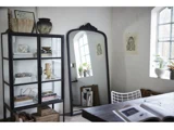 Et hyggeligt, minimalistisk rum med et glasskab med indretning, et stort buet spejl, en stålstol, et skrivebord med en åben bog og lys fra et vindue med planter i karmen.