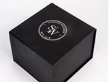 En sort æske med et rundt hvidt logo på toppen, hvor der står Schmidt Watches SW Est. 2018 på en almindelig hvid baggrund.