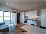 Moderne åbent køkken og opholdsstue med hvide skabe, trægulve, sofa, spisebord og stort vindue med udsigt til kysten.