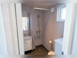 Lille badeværelse med håndvask og spejl til venstre, bruser med forhæng i hjørnet, toiletpapirholder og et lille vindue på højre væg.