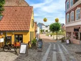 En brostensbelagt gade med gule og murede bygninger, udendørs skiltning, en cykel, træer og runde gule blomsterdekorationer, der hænger ovenover.