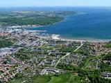 Luftfoto af en kystby med klynger af huse, marina, strand og omkringliggende grønne marker ved siden af en stor mængde blåt vand.