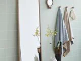 Et badeværelse med et højt spejl, et lille rundt vægspejl, to håndklæder på kroge, en badehandske og en vase med gule blomster på en bordplade mod lysegrønne flisevægge.