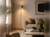 En høj gulvlampe med guldfinish står ved siden af en sofa i neutrale farver og et sofabord af marmor, der er pyntet med bøger og en vase.