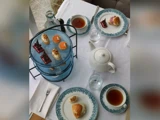 Et afternoon tea-setup med to kopper te, en hvid tekande, scones, kager og forskellige desserter på et blåtonet stativ oven på en hvid dug.