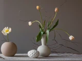 På et bord står tre vaser i neutrale farver med sarte tulipaner og grene, og mellem vaserne er der placeret et hvidt ornament med struktur på en grå baggrund.