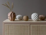Et beige skab med strukturerede vaser og dekorative genstande i neutrale farver står op ad en taupefarvet væg og fremstår i en minimalistisk, moderne stil.