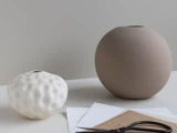To minimalistiske vaser – en lille, struktureret hvid vase og en stor, glat beige vase – står på en hvid overflade ved siden af nogle papirark og en gammel saks.
