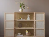 Et minimalistisk træskab med flettede låger, der rummer vaser i neutrale farver, små skulpturer og fuglefigurer. Øverst står en vase med hængende tulipaner.