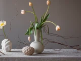 Tre keramiske vaser med blomster og kviste på en struktureret dug. Den ene vase indeholder tulipaner, den anden en enkelt blomst, og foran står et objekt med struktur. Neutral, minimalistisk indretnin