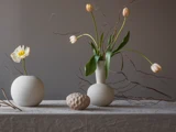 Tre minimalistiske vaser med blomster og kviste står på en struktureret dug foran en grå baggrund; mellem dem ligger der en rund genstand.