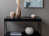 Tre sandfarvede keramiske vaser står på et sort konsolbord; den ene indeholder tørrede blomsterstængler, og på den nederste hylde står en stor hvid vase med bøger.