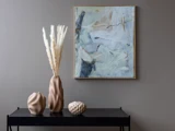 Et sort bord med tre skulpturelle beige vaser og tørret græs står under et abstrakt blåt og beige maleri på en grå væg.