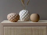 Tre keramiske vaser med struktur – én beige, én hvid og én lysebrun – står på et flettet skab. Den hvide vase er fyldt med tørret græs. En neutral, minimalistisk indretning.