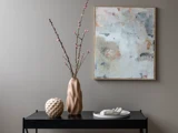 Et sort bord med en bølget vase med lyserøde blomster, en rund skulptur med struktur og et hvidt håndbakke, med et abstrakt maleri på en grå væg bagved.