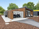Moderne træhus med store vinduer, en åben garage med plads til to golfvogne, omgivet af træer, terrasse og udendørs opholdsområde.