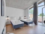 Moderne hotelværelse med to enkeltsenge, hvidt sengetøj, store vinduer med gardiner, trægulv og udsigt til træer og en terrasse udenfor.