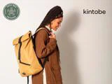 En kvinde med flettet hår smiler, mens hun bærer en stor gul rygsæk og er iført en brun jakke. Kintobe-logoet ses i øverste højre hjørne.
