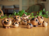 En gruppe små, runde dyrefigurer af træ med store, sorte øjne står på et træbord med grønne planter i baggrunden.