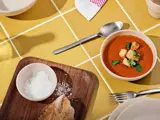 En skål tomatsuppe med croutoner og krydderurter, brød med salt på en træbakke og bestik på et gult flisebord.