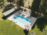 Luftfoto af en rektangulær swimmingpool med liggestole ved poolen, parasoller og et skyggefuldt siddeområde i en græsklædt have omgivet af træer.