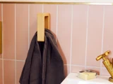 Et mørkt håndklæde hænger på en trækrog mod en lyserød flisebelagt væg ved siden af en vask med sæbe og en messinghane.