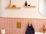 Et moderne badeværelse med lyserøde fliser, guldarmaturer, et håndklæde på en trækrog, hylder med toiletartikler og en del af et spejl, der er synligt.