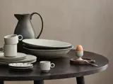 Et sæt keramiske fade, kopper og skåle på et mørkt rundt bord med en kande og et æg i et æggebæger på en træbakke med en ske. Neutrale, jordnære toner hele vejen igennem.