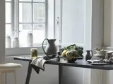 Et køkkenbord med grøntsager, tallerkener, krukker, en kande og et håndklæde nær et vindue med hvide dunke i vindueskarmen.
