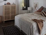 Et hyggeligt soveværelse med et ternet sengetæppe, forskellige puder, et brunt tæppe, et træskab, en sort lampe og et indrammet billede af en abe på en hvid væg beklædt med træpaneler.