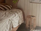Et hyggeligt hjørne i soveværelset med en seng, der er dækket af et stribet beige og hvidt tæppe, en stribet pude og et lille sidebord af rattan med en bog på.