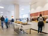 Folk serverer mad for sig selv i et lyst, moderne cafeteria med kurve med frugt og brød på disken.