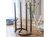 En moderne lysestage i sort metal med tre høje sorte lys på et spisebord i træ dækket med glas, tallerkener og en blå serviet; i baggrunden ses blomster og gardiner.