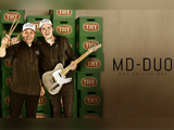 To smilende mænd med matchende MD-DUO-kasketter, der holder trommestikker og en elektrisk guitar, står foran stablede grønne THY-kasser. MD-DUO Det Spiller Bar-teksten står ved siden af dem.