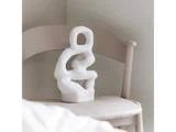 En hvid abstrakt skulptur med afrundede former hviler på en lys beige stol i et blødt oplyst, minimalistisk rum.