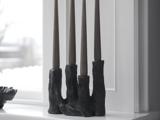 En sort, skulpturel lysestage med fire høje, mørke fyrfadslys står i en hvid vindueskarm foran et vindue.