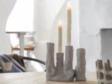 To tændte stearinlys i en abstrakt, grå stenlysestage står på et træbord i et svagt oplyst, minimalistisk rum.