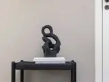 Abstrakt sort skulptur står på et lille bord med to stablede bøger mod en almindelig beige væg i en minimalistisk, moderne indretning.