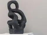 En sort abstrakt skulptur med buede, sammenflettede former og åbne rum, placeret på en stak hvide papirer mod en almindelig grå baggrund.
