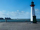 Et fyrtårn står på en brostensbelagt mole ud til havet, med bænke i nærheden og et andet fyrtårn, der kan ses i det fjerne under en blå himmel.