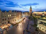 Et livligt bytorv i skumringen med udendørs caféer, gående mennesker, historiske bygninger og et højt klokketårn under en blå himmel.