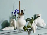 Hvide keramikvaser formet som abstrakte mennesketorsoer og -kroppe rummer blomster, planter og kunstartikler på en pastelblå hylde mod en matchende væg.