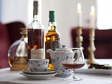 Et bord med spiritusflasker, en tekop, en tekande, et glas med brun væske og to tændte stearinlys i baggrunden.