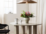 Et rundt beige bord med en vase med rosa og grønne blomster, to keramikkopper, en sort stol og en metallisk hængelampe i et lyst, minimalistisk rum med hvide gardiner.