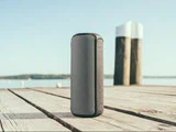 En bærbar Bluetooth-højttaler står på en træbrygge nær roligt vand med to bryggepæle og en klar himmel i baggrunden.