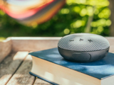 En bærbar Bluetooth-højttaler sidder oven på en lukket bog på en træbakke udendørs, med grønne planter og en farverig hængekøje i den slørede baggrund.